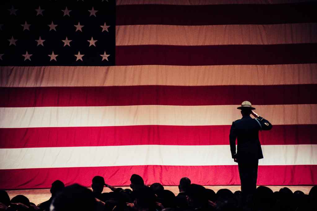 man standing on stage facing an american flag